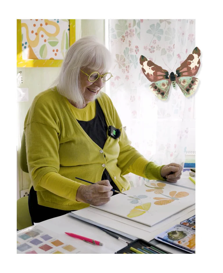 Gudrun Sjödén som sitter vid ett skrivbord och målar fjärilar med akvarellfärg. I bilden finns också en frilagd akvarellfjäril inlagd.