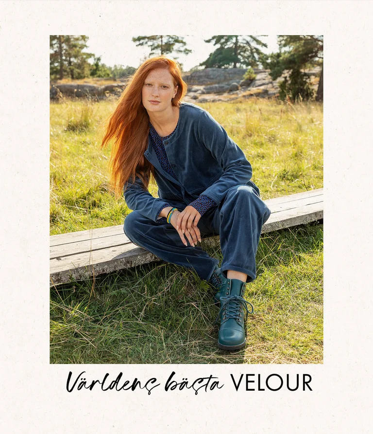 A woman sitting in a green meadow with the forest in the background, wearing blue velour clothing and blue boots from Gudrun Sjödén.