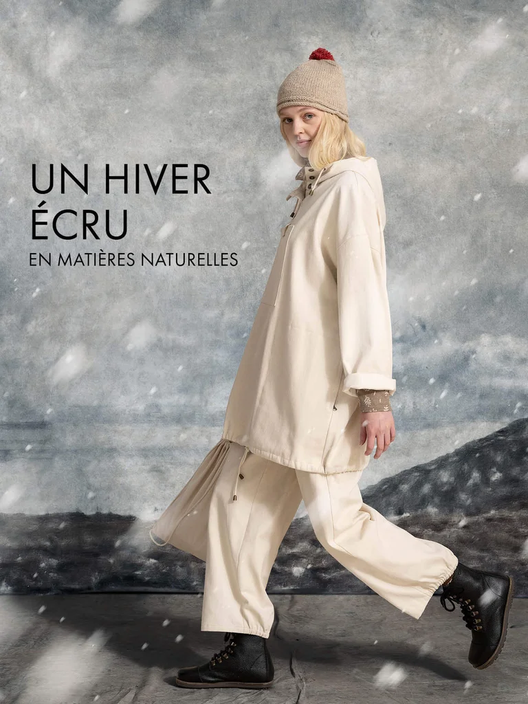 A woman walking while snow falls, wearing a knit hat, a white anorak and a pair of white pants from Gudrun Sjödén.