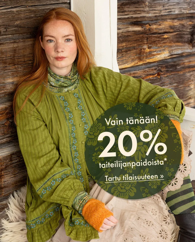 Gudrun Sjödénin vuoden 2025 joulumalliston vihreään kirjailtuun puseroon pukeutunut nainen, joka istuu nojaten puuseinään.