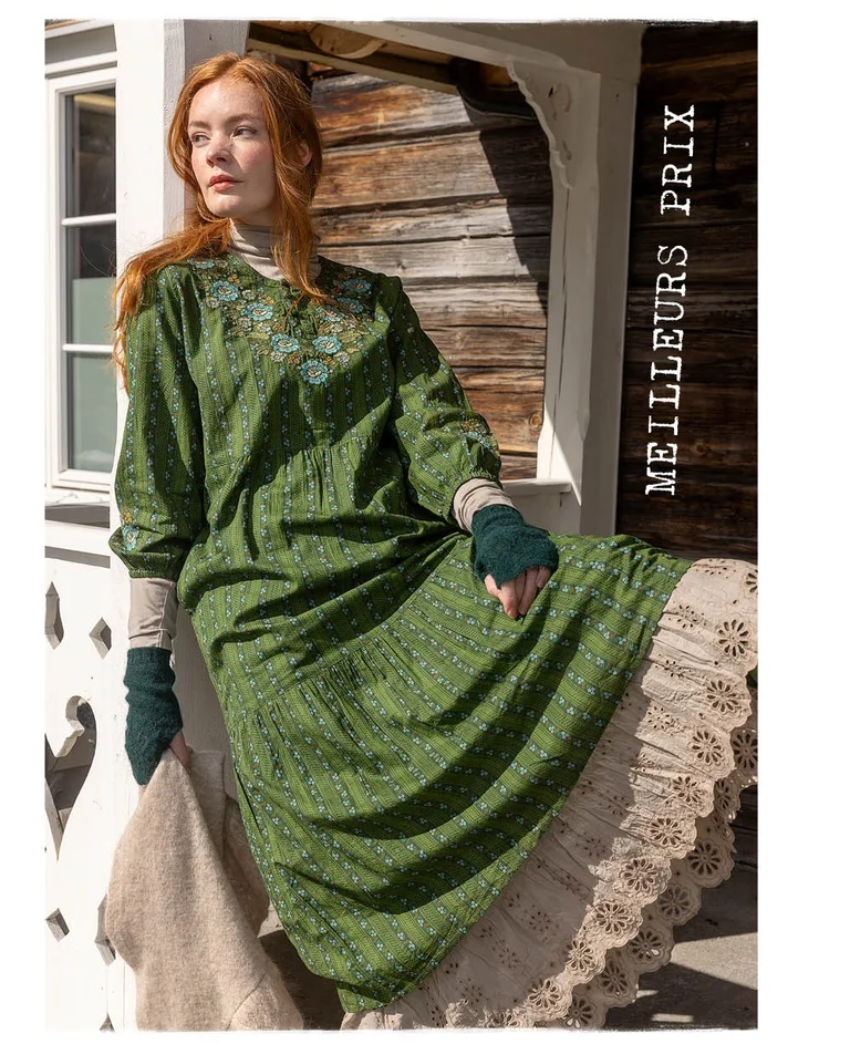 Une femme assise sur le parapet d'un pont et portant des superpositions de vêtements verts et beiges provenant de la collection de Noël 2025 de Gudrun Sjödén.