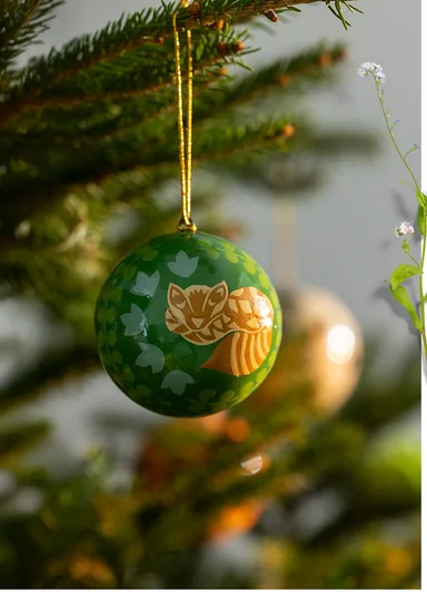 A green bauble with a fox motif from Gudrun Sjödén’s 2025 Christmas Home collection. The bauble is hanging from a fir branch.