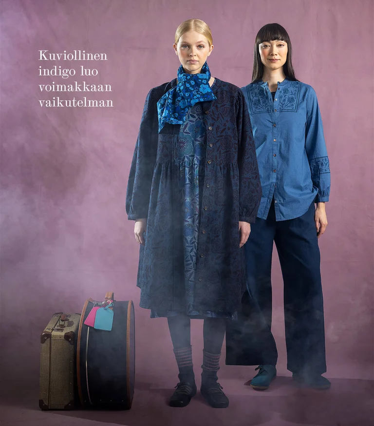 Two women next to each other wearing layers of clothing in shades of blue from Gudrun Sjödén. There are suitcases on the floor next to them.