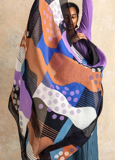 A woman holding up a large, patterned scarf from Gudrun Sjödén.