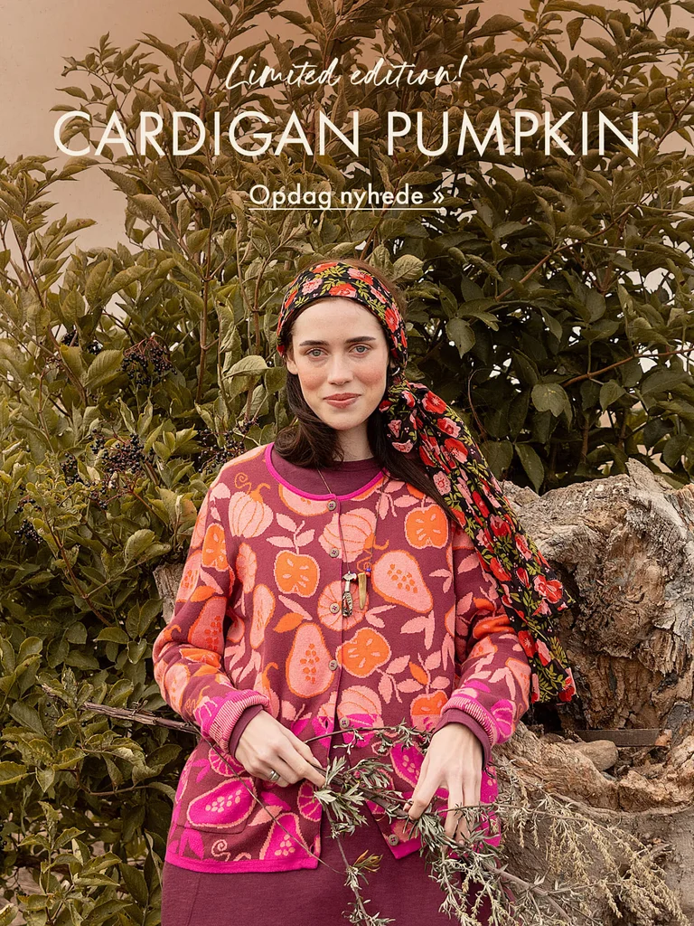 En kvinde foran en frodig busk i et efterårslandskab, iført en mønstret, strikket cardigan fra Gudrun Sjödén med græskar og frugter i orange og rosa.
