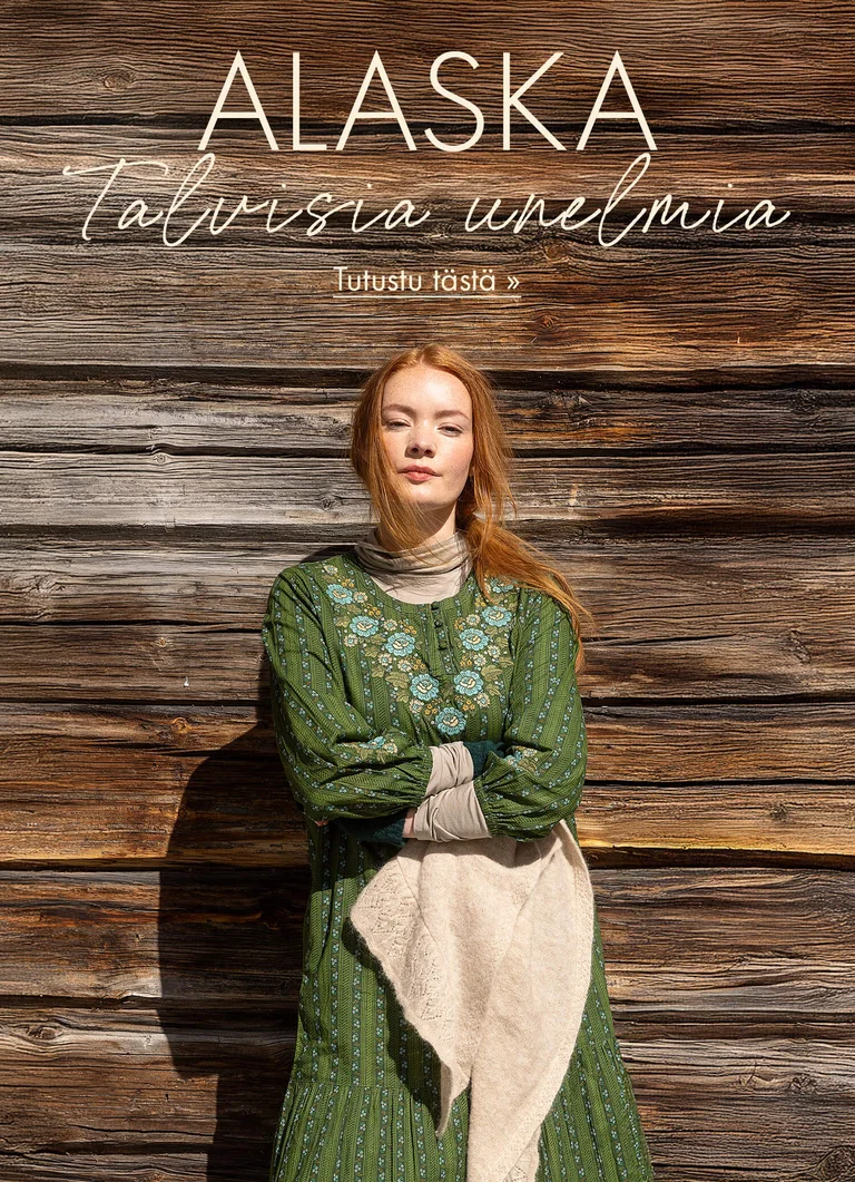 Gudrun Sjödénin vuoden 2025 joulumalliston vihreään kirjailtuun mekkoon pukeutunut nainen, joka seisoo puuseinää vasten käsivarret ristissä.