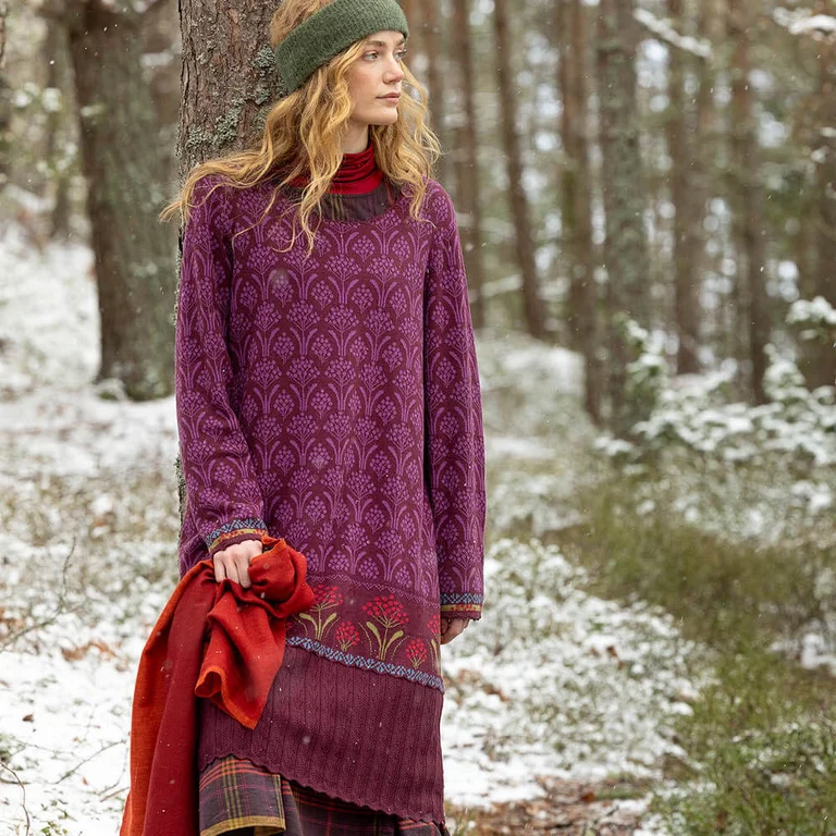 A woman standing in a forest wearing a reddish purple dress from Gudrun Sjödén. 