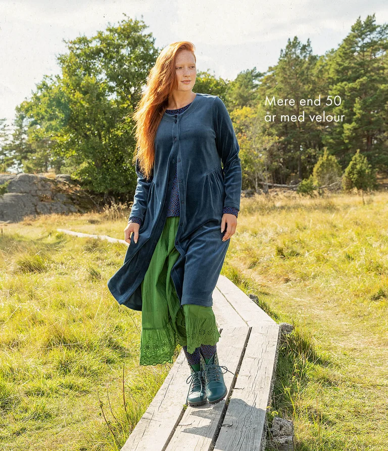 A woman walking on a boardwalk wearing a buttoned, long blue velour dress from Gudrun Sjödén.