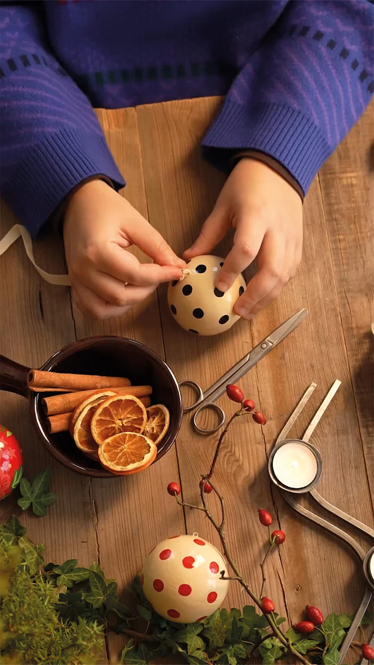 Een video waarin handen handgeschilderde kerstballen en andere hangers van Gudrun Sjödén versieren in een warme, creatieve omgeving met gedroogde sinaasappels en linten.