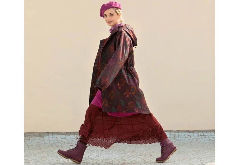 A woman wearing a patterned longline jacket, a burgundy lace underskirt, boots and a beret from Gudrun Sjödén.