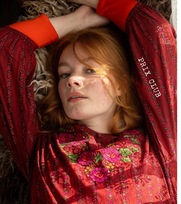 Une femme allongée portant une robe brodée rouge de la collection de Noël 2025 de Gudrun Sjödén. 