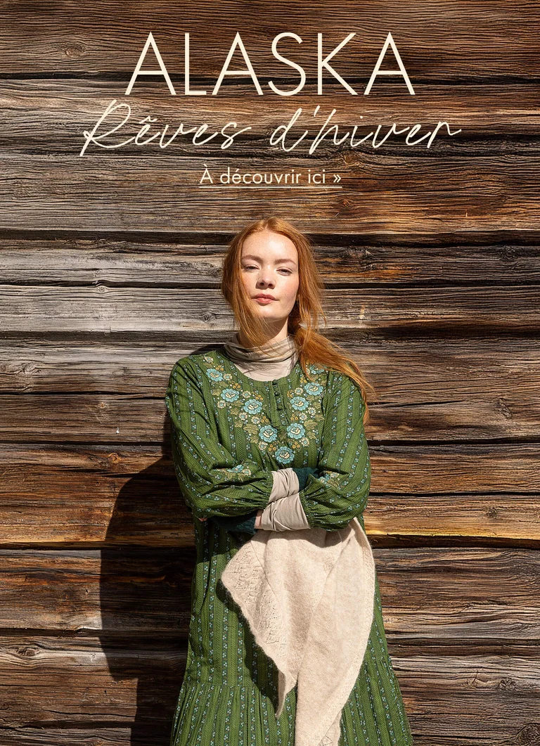 Une femme se tenant contre un mur en bois avec les bras croisés et portant une robe verte brodée provenant de la collection de Noël 2025 de Gudrun Sjödén.