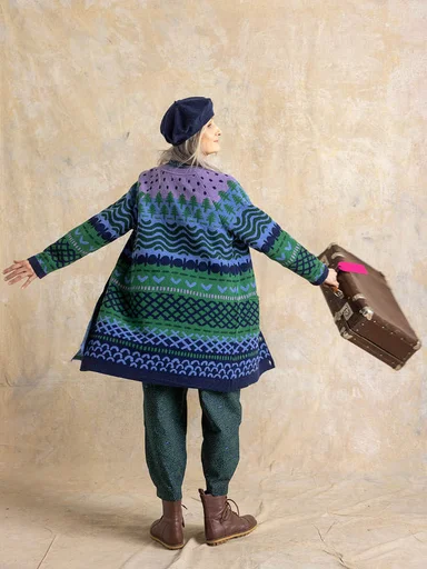 A woman with her back to the camera wearing a patterned long cardigan and pants from Gudrun Sjödén. She holds a suitcase in her hand.