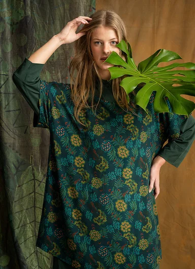 A woman wearing a patterned green top with a branch with leaves along one side of her face.