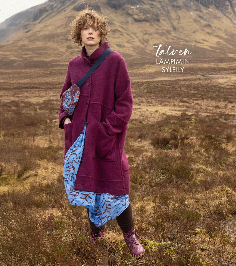 A woman in a mountain landscape wearing a burgundy wool coat, a light blue patterned dress and a crossbody bag from Gudrun Sjödén.