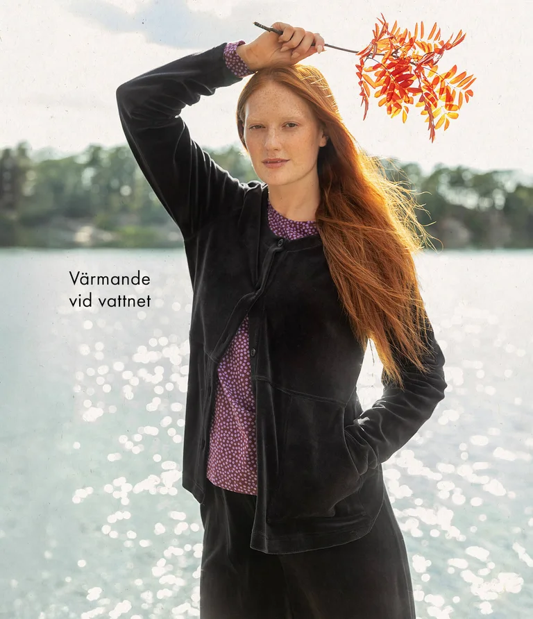A woman in front of water wearing a black velour cardigan with matching velour pants from Gudrun Sjödén. She is holding a branch from a mountain ash tree in her hand.