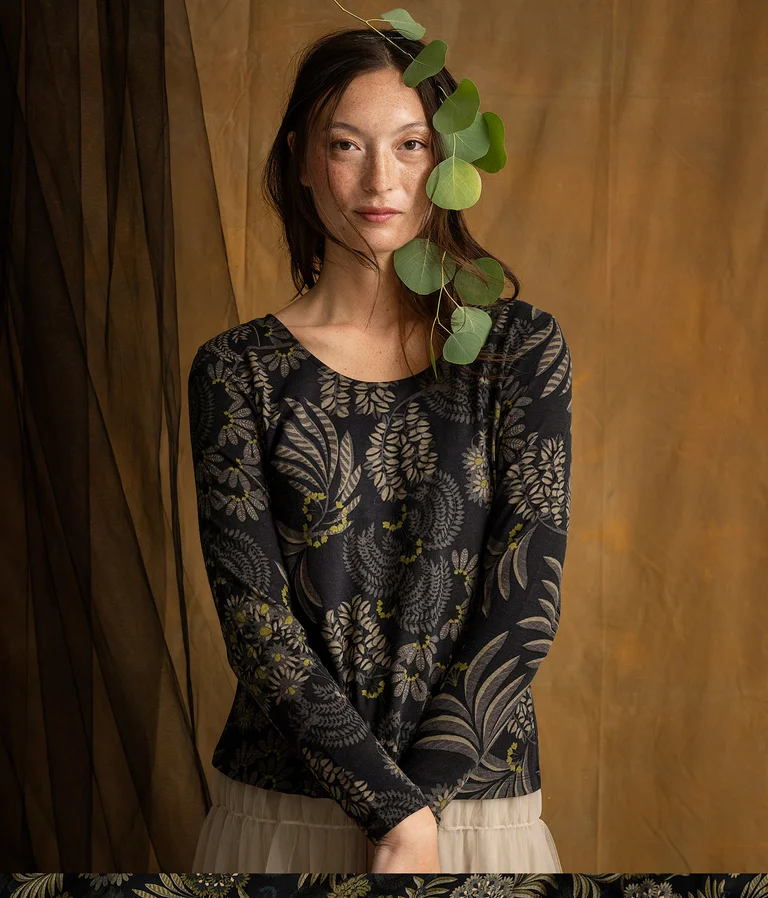 A woman wearing a patterned black top with a branch with leaves along one side of her face.