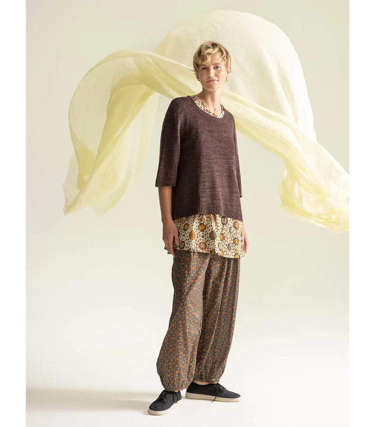 Eine Frau, die einen braunen Pullover, eine gemusterte Tunika und Hosen von Gudrun Sjödén trägt.
