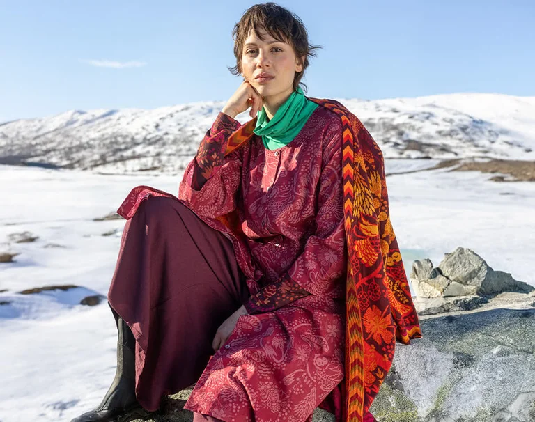 En kvinne som sitter på en stein i et snølandskap kledd i lag på lag med klær fra Gudrun Sjödéns julekolleksjon 2025.  