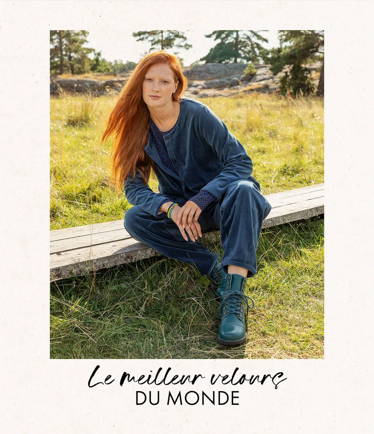 A woman sitting in a green meadow with the forest in the background, wearing blue velour clothing and blue boots from Gudrun Sjödén.