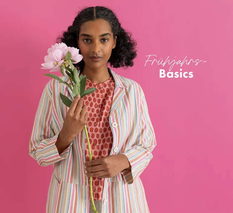 Eine Frau vor rosafarbenem Hintergrund, in Basics aus Gudrun Sjödéns Frühjahrskollektion 2026. In der Hand eine rosa Blume. 