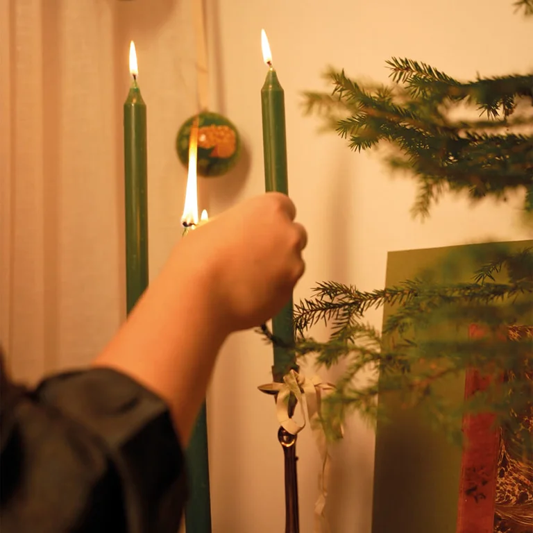 Hands lighting green candles in a candle holder decorated with sprays of spruce.