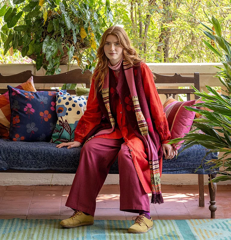 A woman wearing an outfit of reds and pinks, seated among patterned cushions from Gudrun Sjödén.