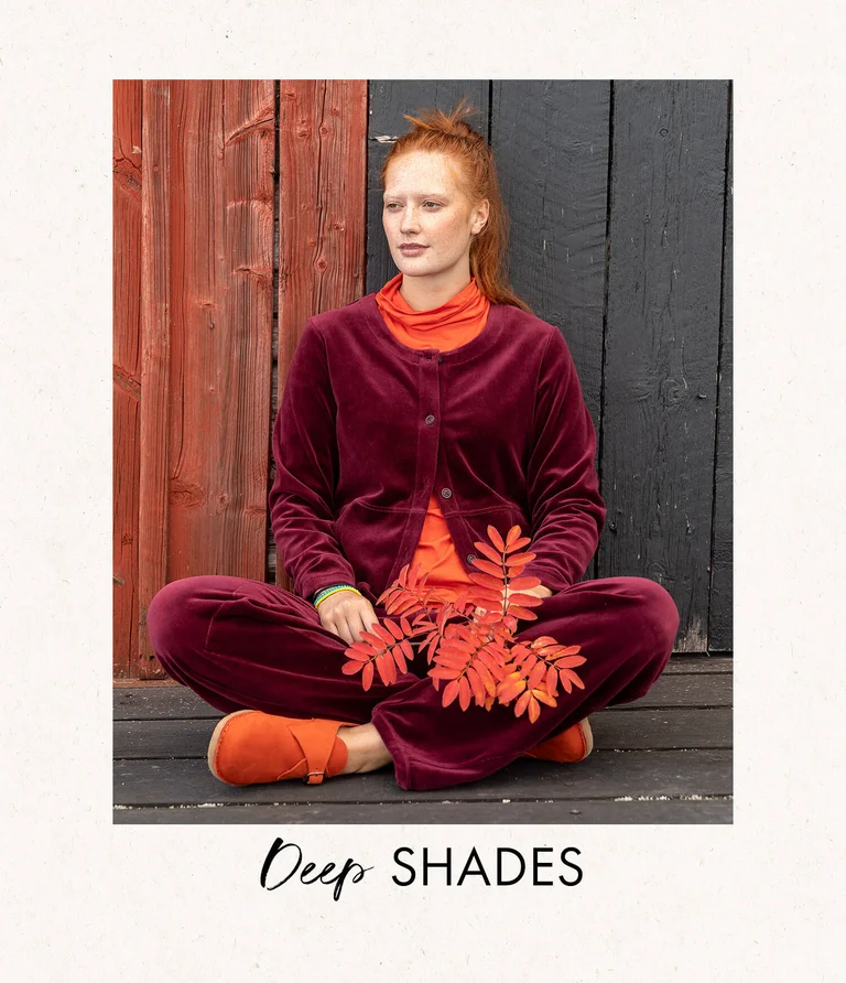 A woman sitting outdoors wearing a red velour cardigan with matching velour pants from Gudrun Sjödén. She is holding a branch from a mountain ash tree in her hand.