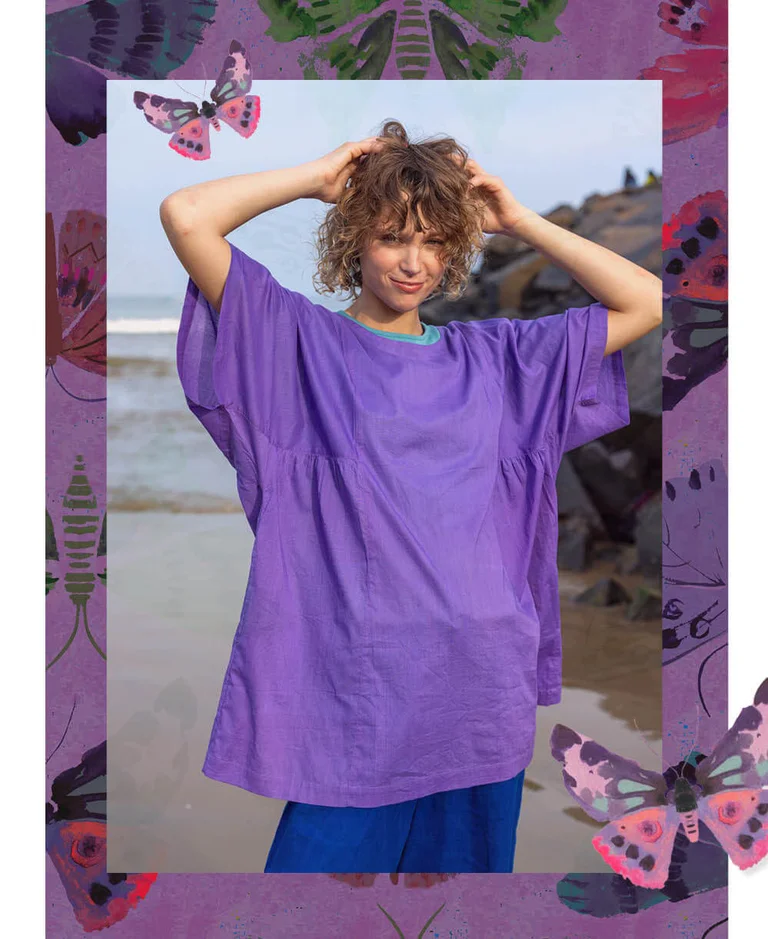 A woman standing on a beach, hands on her head, wearing a purple tunic from Gudrun Sjödén. With a purple frame and standout watercolour butterflies. 