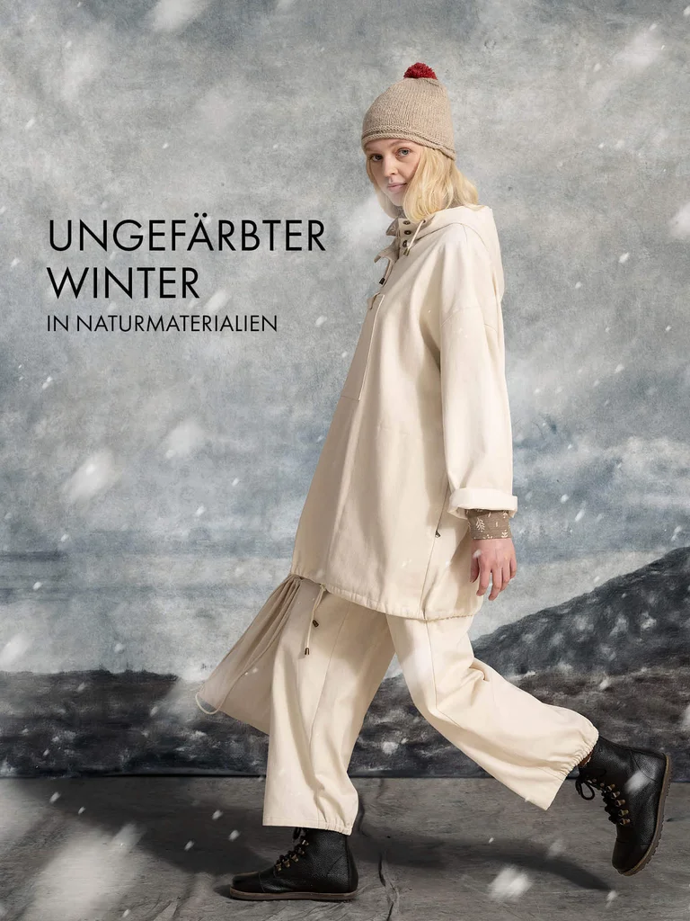 A woman walking while snow falls, wearing a knit hat, a white anorak and a pair of white pants from Gudrun Sjödén.