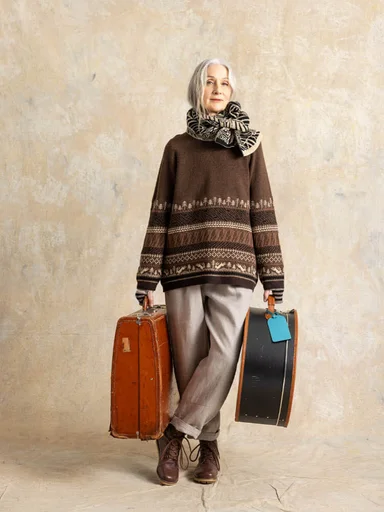 A woman wearing a scarf, pants and a patterned brown sweater from Gudrun Sjödén. She holds suitcases in her hands.