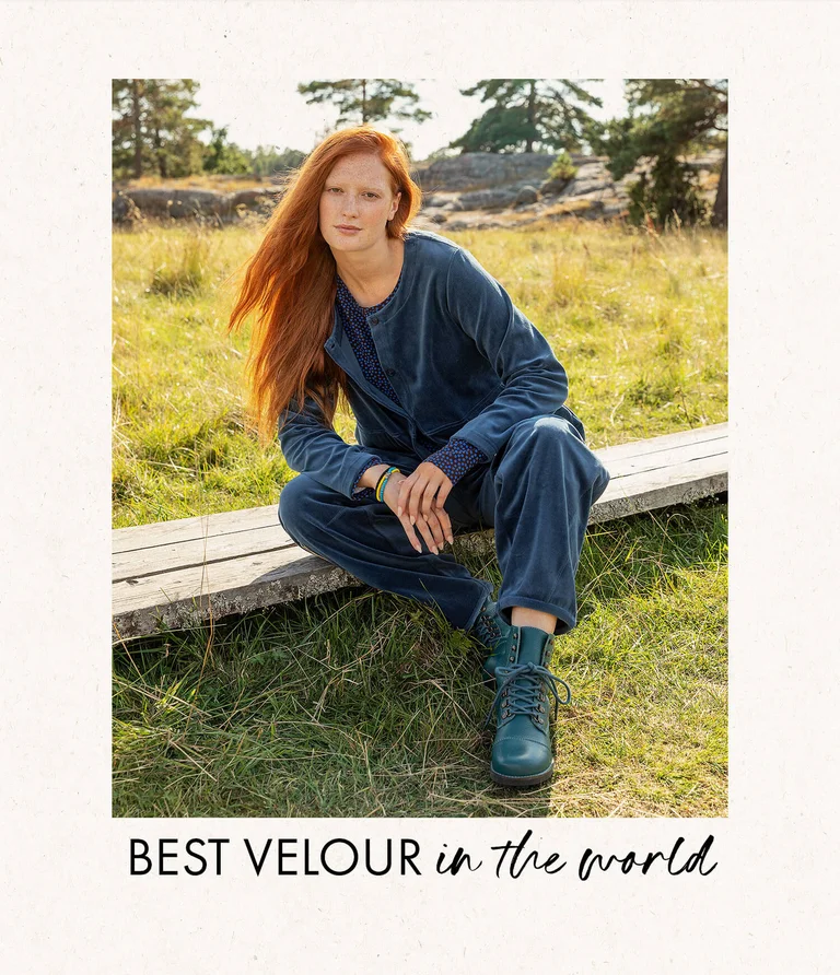 A woman sitting in a green meadow with the forest in the background, wearing blue velour clothing and blue boots from Gudrun Sjödén.