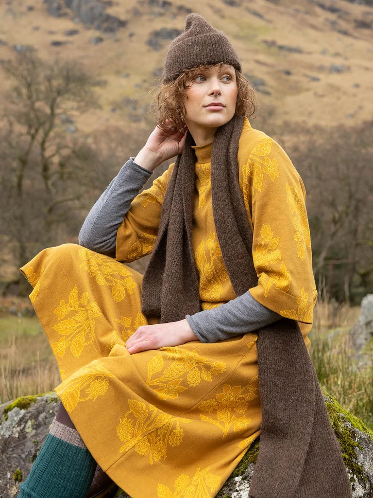 A woman sitting and wearing a yellow dress and brown knit accessories from Gudrun Sjödén.