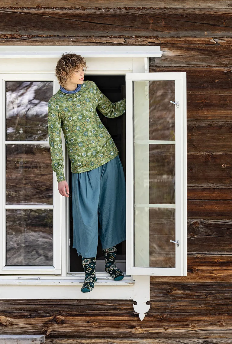 A picture of three women standing next to a timber house wearing clothes from Gudrun Sjödén’s 2025 Christmas collection.