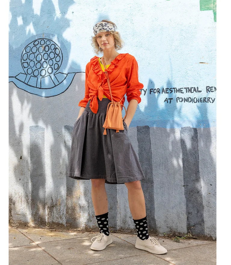 A woman standing next to a wall wearing an orange ruffle blouse paired with a jeans skirt from Gudrun Sjödén.