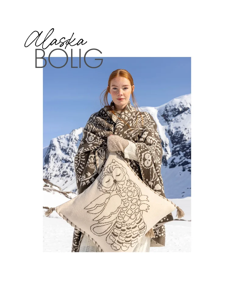 A woman standing in a snowy landscape wrapped in a blanket, holding a cushion, from Gudrun Sjödén’s 2025 Christmas Home collection. 