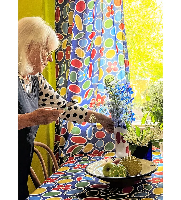 Gudrun Sjödén is arranging flowers at a kitchen table with a cobalt blue floral tablecloth from Gudrun Sjödén. Behind her is a curtain with the same pattern hanging in front of a sunny window. She is wearing a black-and-white dotted top from Gudrun Sjödén.