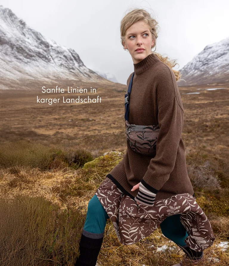 A woman walking in a mountain setting wearing a brown knit tunic and a patterned crossbody bag from Gudrun Sjödén.