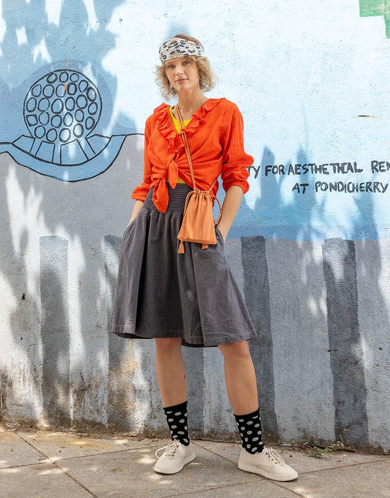 A woman standing next to a wall wearing an orange ruffle blouse paired with a jeans skirt from Gudrun Sjödén.