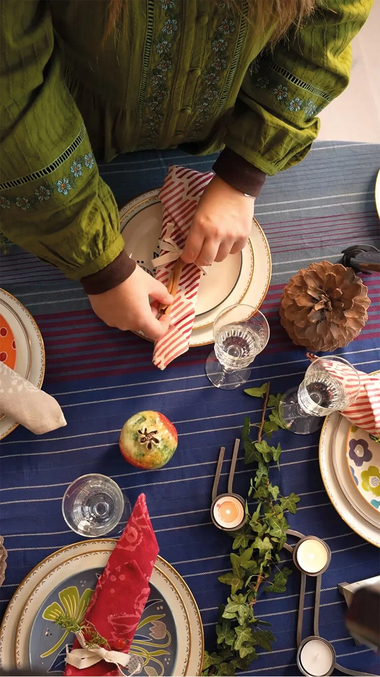 Handen die de tafel feestelijk dekken met woontextiel van Gudrun Sjödén, vintage servies en groene decoraties.
