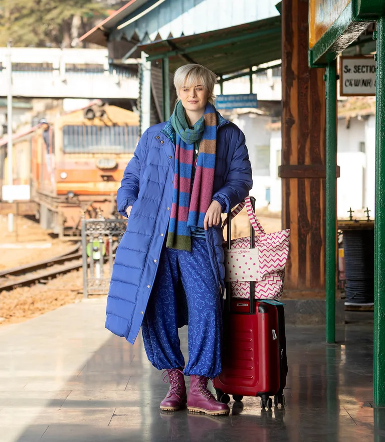 A woman by a suitcase in a train station, wearing a long blue down coat over layers of clothing from Gudrun Sjödén.