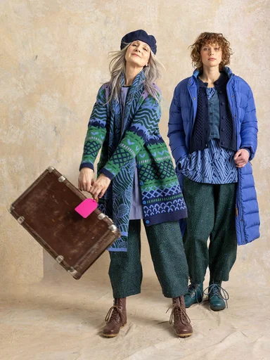 Two women next to each other wearing layers of clothes in shades of blue and green from Gudrun Sjödén. One with a travel bag swinging from her hand.