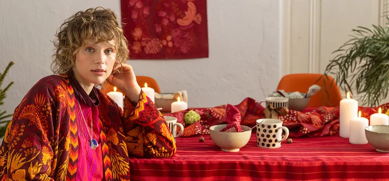 A woman sitting next to a table with lit candles, wearing red and pink clothes from Gudrun Sjödén’s 2025 Christmas collection.