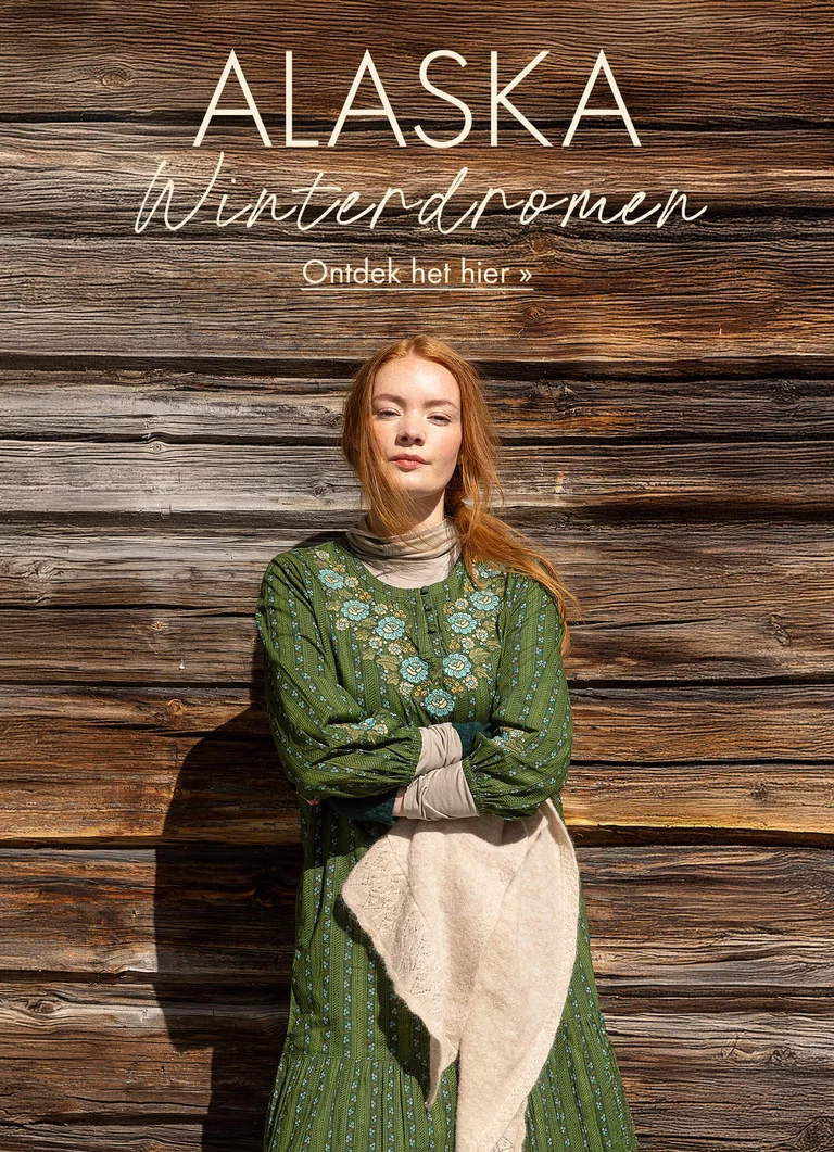 Een vrouw die met gekruiste armen tegen een houten wand staat en een groene jurk met borduursel uit de kerstcollectie 2025 van Gudrun Sjödén draagt. 