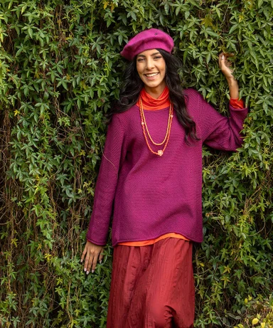 A woman standing against a verdant background wearing layers of pink and red clothes from Gudrun Sjödén.