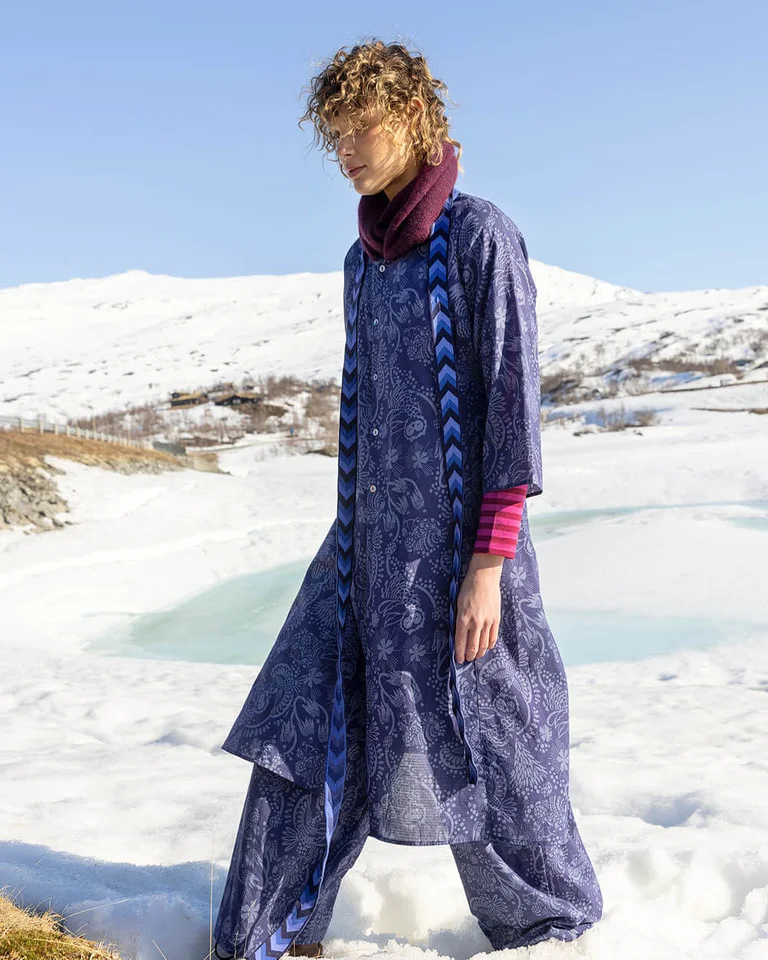 A woman walking in a snowy landscape wearing layers of clothes from Gudrun Sjödén’s 2025 Christmas collection.