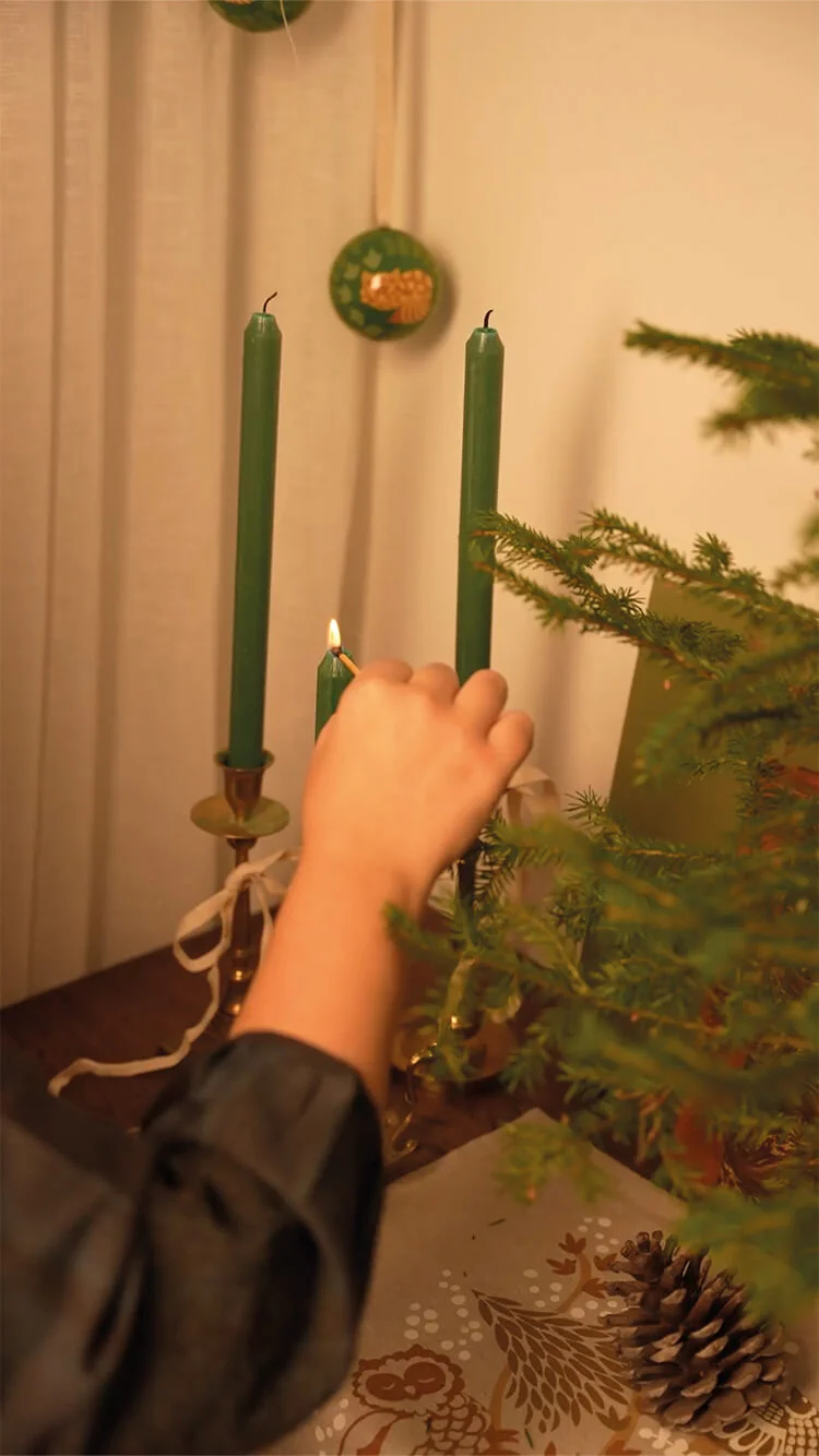 Handen die groene kaarsen in drie kandelaars aansteken. Aan het einde hangen de handen een rode kerstbal in dennentakken bij de kaarsen. 