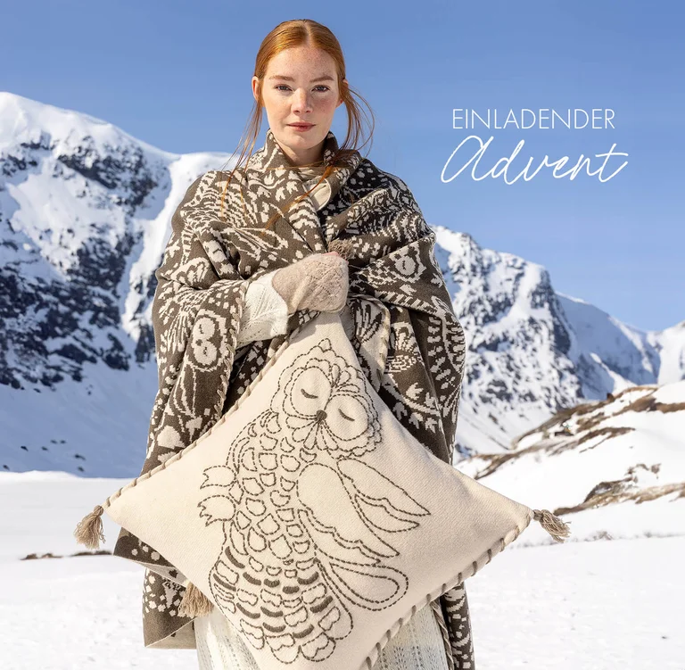 Eine Frau in verschneiter Landschaft, in der Hand ein Kissen und umhüllt von einem grauen Plaid. Produkte aus Gudrun Sjödéns weihnachtlicher Zuhause-Kollektion 2025. 