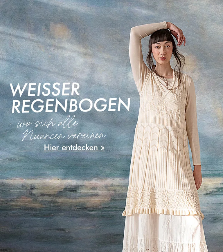 Eine Frau im weißen Lagenlook von Gudrun Sjödén, vor einem Hintergrund, der einem abstrakten Ölgemälde in blaugrauen Nuancen gleicht.