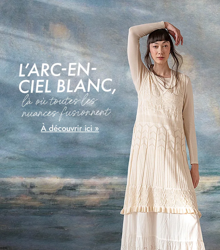 Une femme portant des vêtements blancs signés Gudrun Sjödén superposés devant une peinture abstraite aux nuances bleu gris. 